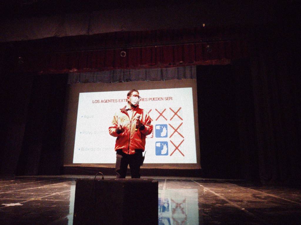 Un presentador con chaqueta roja y mascarilla habla frente a un proyector en un auditorio, mostrando diapositivas sobre agentes externos de riesgo.